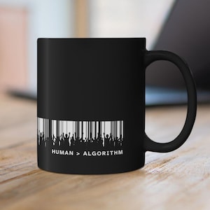 May include: Black ceramic mug with a white graphic. The graphic features a barcode design with silhouetted figures and the text "HUMAN > ALGORITHM". The mug has a curved handle and is set against a blurred background.