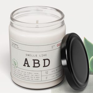 May include: A white scented candle in a clear glass jar with a black lid. The label reads "SMELLS LIKE ABD" with additional text. The candle has a burn time of 50-60 hours and weighs 9 oz.
