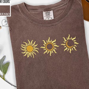Op de afbeelding: Espresso-kleurig T-shirt met drie geborduurde gele zonontwerpen. Het shirt heeft een ronde hals en korte mouwen. De merknaam "Comfort Colors" is zichtbaar in de linkerbovenhoek.
