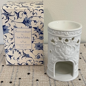 May include: White ceramic oil diffuser with a floral design and a cutout in the center. The diffuser is packaged in a box with a blue and white floral pattern. The box reads "Surroundings by Diane von Furstenberg Fragrant Home Diffuser".