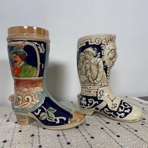 May include: Two ceramic boot-shaped mugs. One is blue, beige, and brown with a painted scene of a man in a green hat. The other is beige with a relief of a couple and a grotesque face. Both have decorative patterns.
