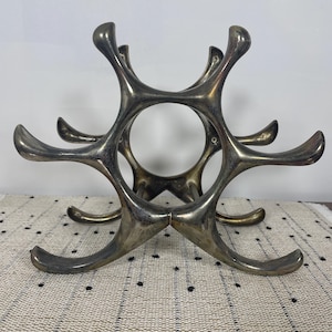 May include: A silver-toned metal wine rack with a unique, abstract design. The rack features a central circular opening surrounded by curved arms, each designed to hold a bottle. The rack is approximately 12 inches wide and 8 inches tall.
