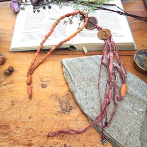 May include: A necklace featuring a strand of orange cylindrical beads connected to a circular wooden pendant. The pendant is attached to several long, frayed strips of dusty pink fabric, accented with small black and white striped beads.
