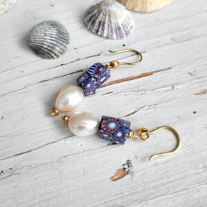 May include: A pair of drop earrings featuring gold-tone hooks, each with a cylindrical bead displaying a blue, white, and red floral-like pattern, followed by a single off-white, irregular-shaped pearl bead, finished with a small gold-tone bead.