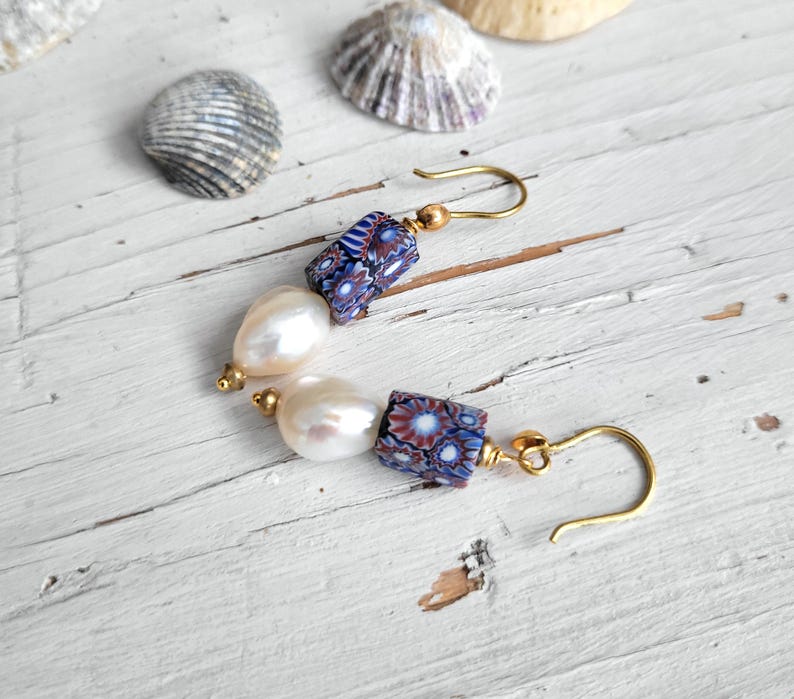 May include: A pair of drop earrings featuring gold-tone hooks, each with a cylindrical bead displaying a blue, white, and red floral-like pattern, followed by a single off-white, irregular-shaped pearl bead, finished with a small gold-tone bead.