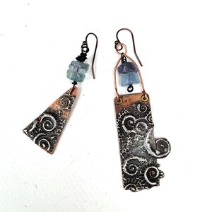 May include: Artisan earrings with a unique design. One earring is a copper triangle with a swirling pattern and blue-green beads. The other is a copper rectangle with a similar pattern and blue-purple beads. Both have copper ear wires.