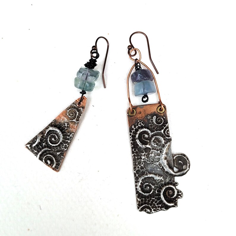May include: Artisan earrings with a unique design. One earring is a copper triangle with a swirling pattern and blue-green beads. The other is a copper rectangle with a similar pattern and blue-purple beads. Both have copper ear wires.
