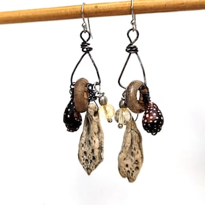May include: A pair of dangle earrings featuring dark wire frames, each adorned with a textured tan ring, a porous beige stone pendant, a small clear yellow bead, and a dark brown shell with white spots, suspended from silver-toned hooks.