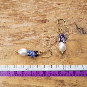 May include: A pair of drop earrings featuring white teardrop-shaped pearls and rectangular millefiori glass beads with blue, red, and white floral patterns, suspended from gold-tone hooks, shown next to a ruler measuring approximately 2.5 inches.