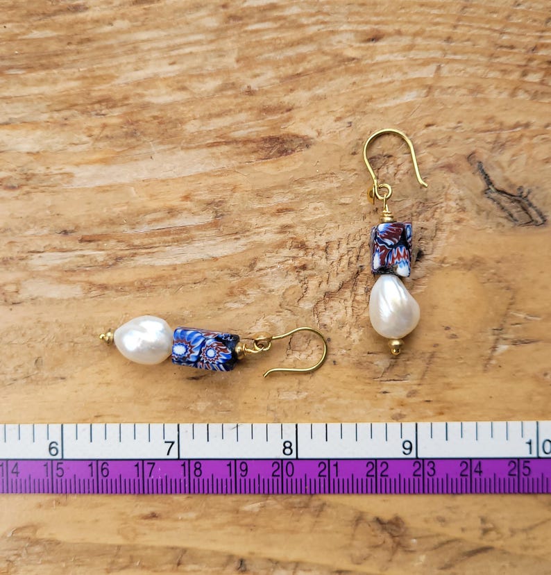May include: A pair of drop earrings featuring white teardrop-shaped pearls and rectangular millefiori glass beads with blue, red, and white floral patterns, suspended from gold-tone hooks, shown next to a ruler measuring approximately 2.5 inches.