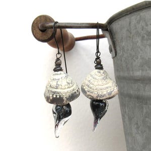 May include: A pair of dangle earrings featuring textured, off-white and grey ceramic beads with a rounded, shell-like shape. Each earring is suspended from a dark metal wire hook and includes a smooth, teardrop-shaped black glass bead at the base.