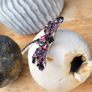 May include: A dragonfly-shaped brooch crafted from silver-toned metal, featuring an array of faceted purple and dark violet crystals. The wings and body are densely encrusted with these shimmering stones, creating a detailed insect design.