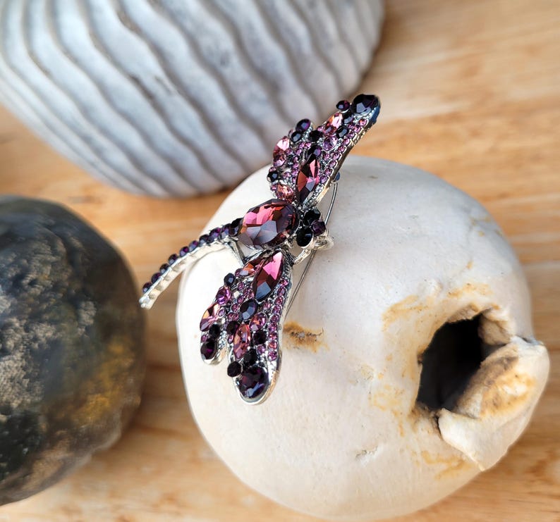 May include: A dragonfly-shaped brooch crafted from silver-toned metal, featuring an array of faceted purple and dark violet crystals. The wings and body are densely encrusted with these shimmering stones, creating a detailed insect design.