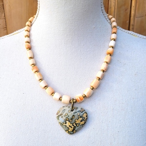 Può includere: Una collana con un ciondolo a forma di cuore. Il ciondolo è un mix di grigio, giallo e marrone. La collana ha perline color pesca con distanziatori color oro. La collana è esposta su un manichino bianco.