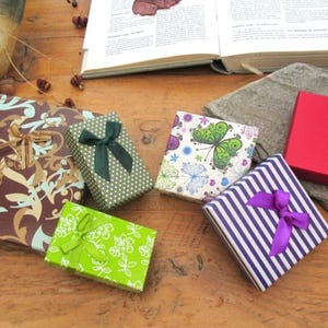 May include: A collection of six square and rectangular gift boxes featuring various designs: a brown box with gold swirls, a green dotted box with a dark green bow, a bright green floral box, a white butterfly print box, a purple striped box with a purple bow, and a red box with two silver hearts.