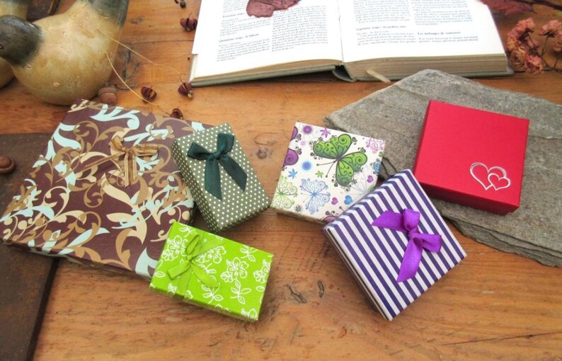 May include: A collection of six square and rectangular gift boxes featuring various designs: a brown box with gold swirls, a green dotted box with a dark green bow, a bright green floral box, a white butterfly print box, a purple striped box with a purple bow, and a red box with two silver hearts.