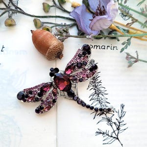 May include: A dragonfly brooch featuring a large, dark purple oval crystal body and wings encrusted with smaller purple and black rhinestones set in a silver-toned metal frame. The insect-shaped pin displays intricate detailing across its wings.