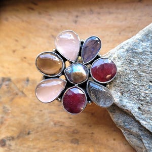 Op de afbeelding: Een zilveren ring met een bloemachtig ontwerp. De ring heeft een centrale iriserende steen, omringd door druppelvormige edelstenen in roze, bruin, paars en rood. De ring ligt op een houten oppervlak.