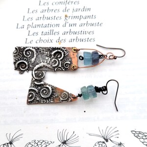 May include: Artisan earrings with a mixed metal design. The earrings feature a silver-tone, textured metal with a copper-tone accent. Each earring has a unique shape and is adorned with blue-green beads. The background includes French text.