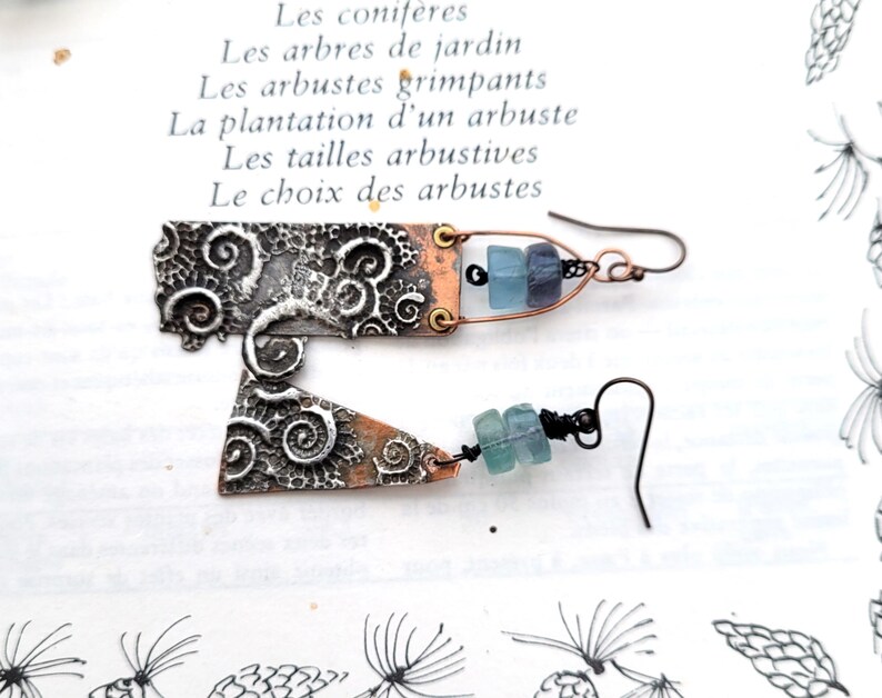 May include: Artisan earrings with a mixed metal design. The earrings feature a silver-tone, textured metal with a copper-tone accent. Each earring has a unique shape and is adorned with blue-green beads. The background includes French text.