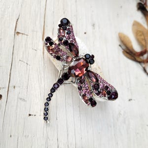 May include: A dragonfly brooch crafted from silver-toned metal, featuring a large oval-cut dark red central stone and wings encrusted with a mix of deep purple, light pink, and dark red rhinestones, with a slender tail lined with small black stones.