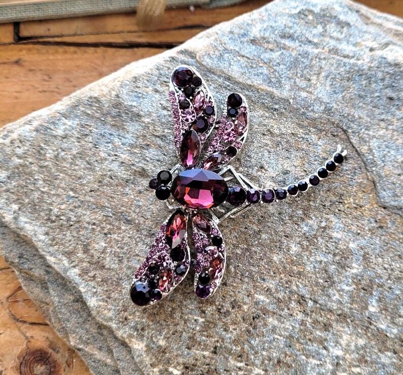May include: A dragonfly brooch featuring a large oval purple crystal body surrounded by smaller dark purple and light pink rhinestones set in a silver-toned metal frame, with a long tail accented by a row of small round black crystals.