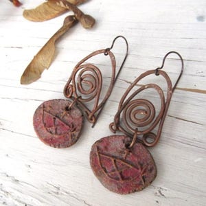 May include: A pair of dangle earrings featuring copper wire formed into a spiral and arch shape, connected to a round, textured, reddish-brown clay pendant with an etched geometric design. The earrings have dark metal hooks for attachment.