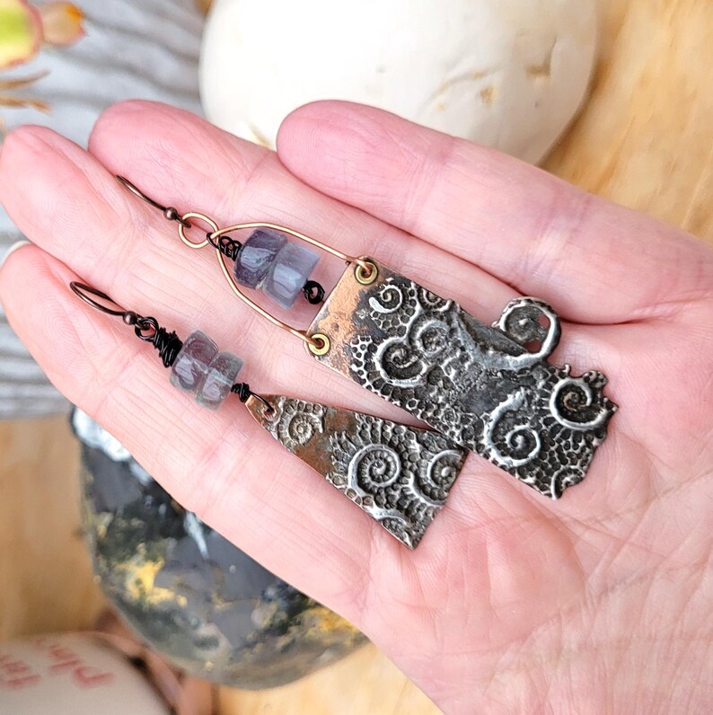 May include: Artisan-made earrings with a unique design. The earrings feature a combination of copper and silver-toned metal, with intricate spiral and swirl patterns. Each earring has a wire hook and is adorned with small, light purple beads.