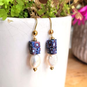 May include: A pair of drop earrings featuring gold-tone hooks, small gold beads, square-shaped beads with a blue, red, and white floral pattern, and teardrop-shaped white pearls at the base. The earrings hang from gold-tone metal wires.