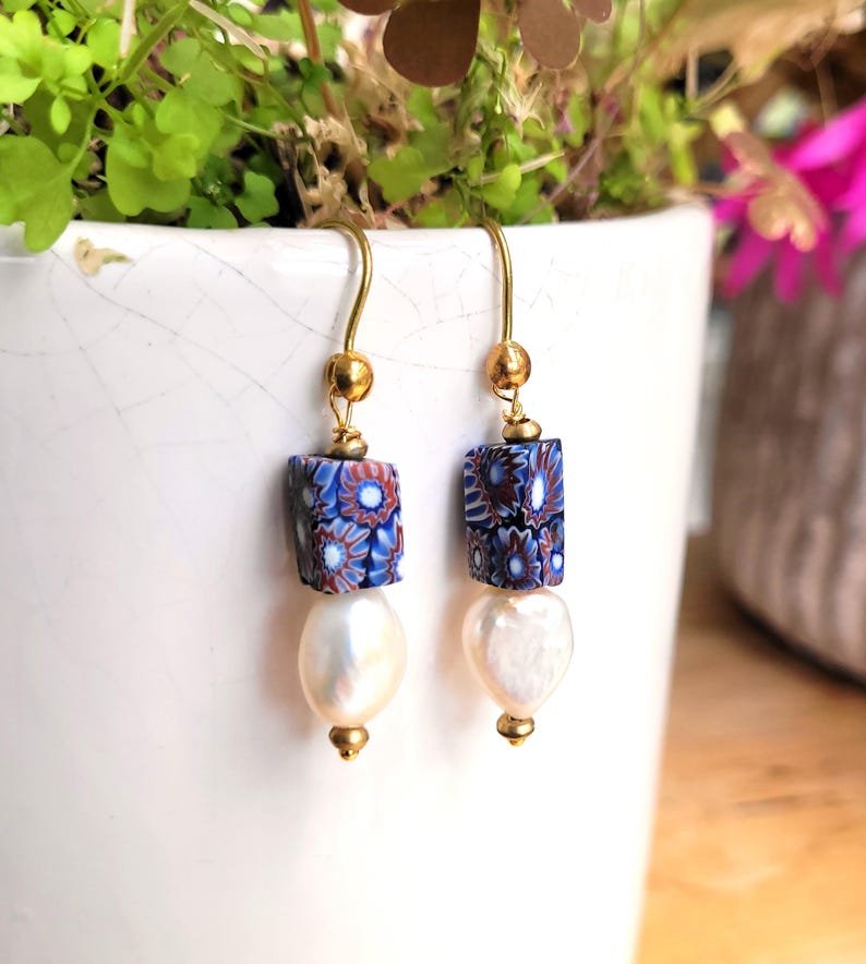 May include: A pair of drop earrings featuring gold-tone hooks, small gold beads, square-shaped beads with a blue, red, and white floral pattern, and teardrop-shaped white pearls at the base. The earrings hang from gold-tone metal wires.