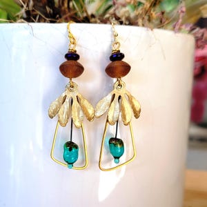 Peut inclure: Une paire de boucles d'oreilles pendantes avec des breloques en métal doré en forme de feuilles suspendues au-dessus d'un cadre en fil trapézoïdal. Chaque boucle comprend une perle violet foncé, une perle brune texturée et une perle en verre turquoise.
