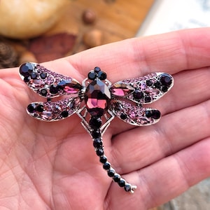 May include: A dragonfly brooch featuring a silver-tone metal frame encrusted with various shades of purple and dark violet crystals. The central body is composed of a large oval purple stone, while the wings are detailed with smaller faceted gems.