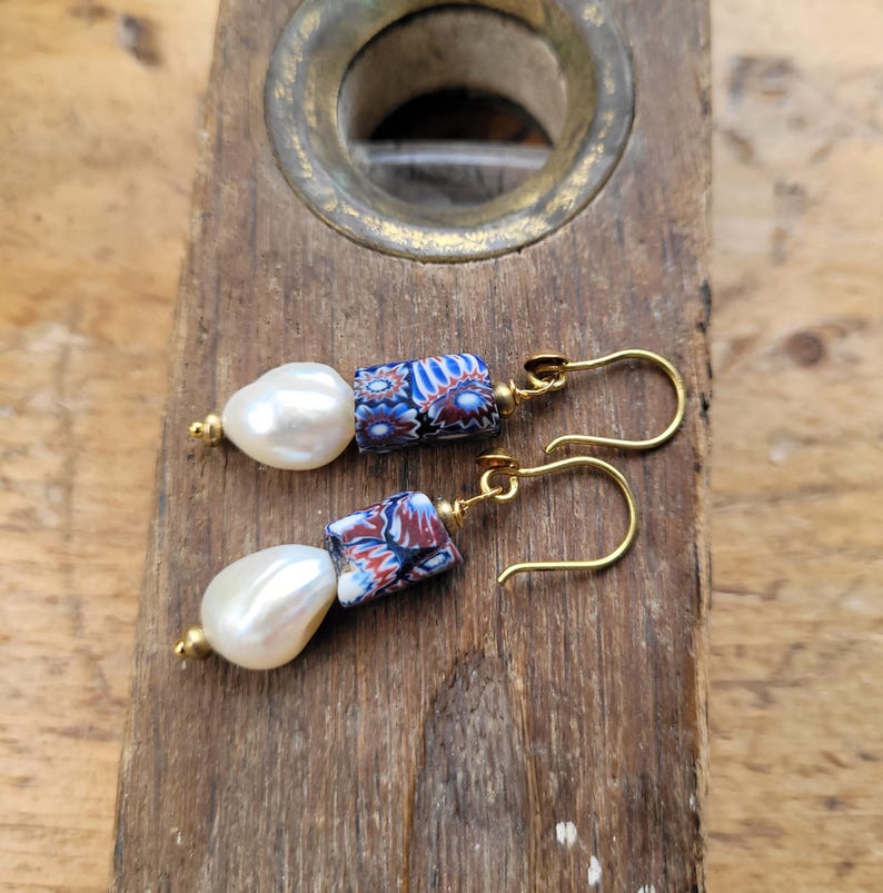 May include: A pair of drop earrings featuring white baroque pearls paired with cylindrical millefiori glass beads in blue, red, and white. The beads are accented with small gold-toned metal spacers and suspended from gold-toned metal ear wires.