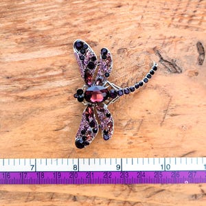 May include: A dragonfly brooch featuring a large oval purple center stone, accented with smaller purple and black crystals set in a silver-tone metal frame. A measuring tape is visible below, showing the brooch spans approximately 2.5 inches.