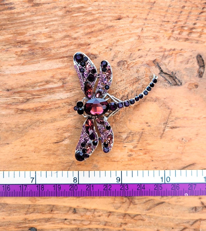 May include: A dragonfly brooch featuring a large oval purple center stone, accented with smaller purple and black crystals set in a silver-tone metal frame. A measuring tape is visible below, showing the brooch spans approximately 2.5 inches.