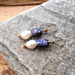 May include: A pair of drop earrings featuring white teardrop-shaped pearls paired with cylindrical beads. The beads display a multi-colored floral pattern in shades of blue, red, and white. The components are connected by gold-toned metal findings.