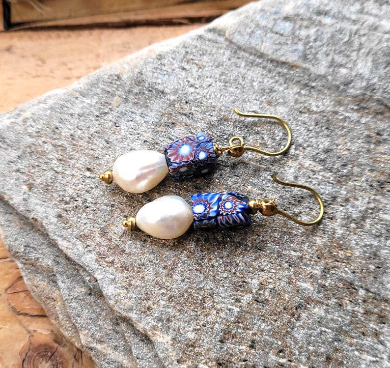 May include: A pair of drop earrings featuring white teardrop-shaped pearls paired with cylindrical beads. The beads display a multi-colored floral pattern in shades of blue, red, and white. The components are connected by gold-toned metal findings.