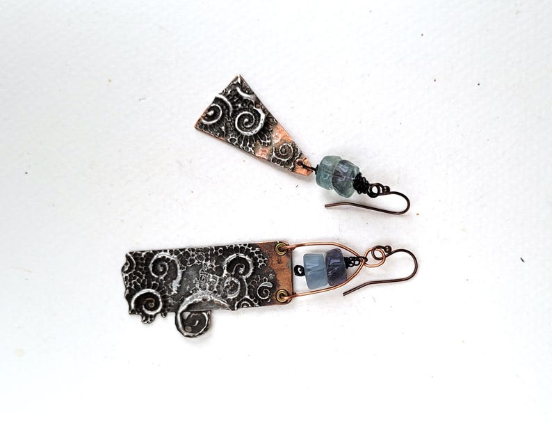 May include: Artisan earrings with a unique design. The earrings feature textured metalwork with swirling patterns and copper accents. Each earring has a dangling element with blue-green and purple beads, suspended from a copper wire hook.
