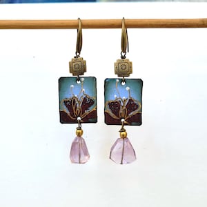 May include: A pair of dangle earrings featuring rectangular metal plaques with a blue and dark red floral enamel design. Each earring includes a square brass accent piece, a gold-toned bead, and a faceted, translucent light purple stone drop.