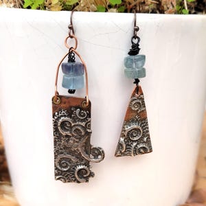 May include: Handmade artisan earrings. One earring is rectangular, the other is triangular, both with a textured, swirling pattern. Each earring features blue-green gemstone beads and copper-colored metal accents. The earrings hang from a simple hook.