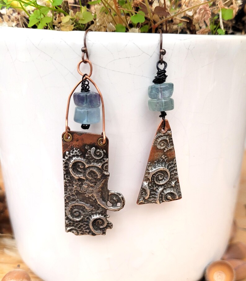 May include: Handmade artisan earrings. One earring is rectangular, the other is triangular, both with a textured, swirling pattern. Each earring features blue-green gemstone beads and copper-colored metal accents. The earrings hang from a simple hook.