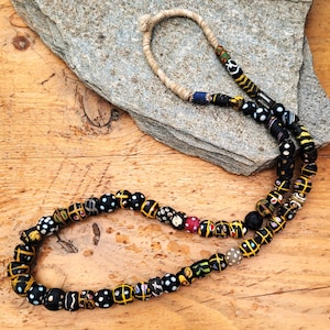 Peut inclure: Un long collier composé de nombreuses perles noires peintes à la main avec divers motifs colorés, notamment des croix jaunes, des points blancs et des accents rouges. Les perles sont enfilées sur une cordelette en fibre beige clair.