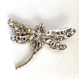 May include: A silver-toned dragonfly brooch featuring a textured, metallic finish on the wings and body. The reverse side displays a horizontal pin mechanism with a secure clasp, showcasing the intricate, openwork metal construction of the insect.