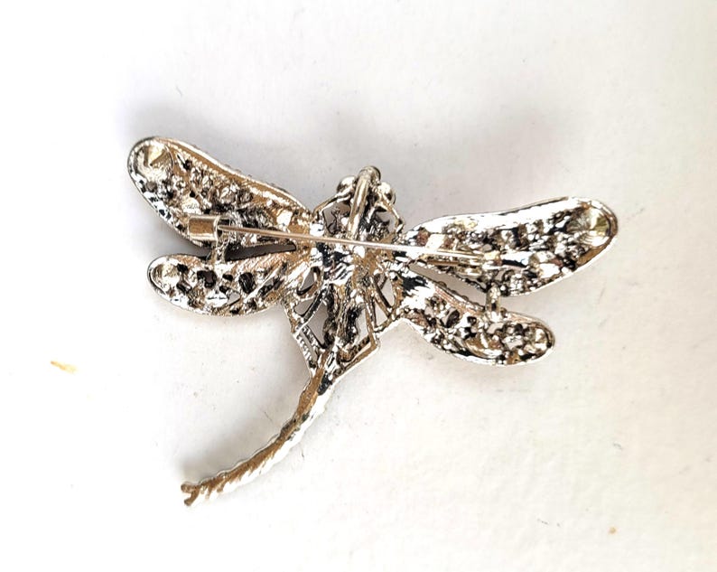 May include: A silver-toned dragonfly brooch featuring a textured, metallic finish on the wings and body. The reverse side displays a horizontal pin mechanism with a secure clasp, showcasing the intricate, openwork metal construction of the insect.