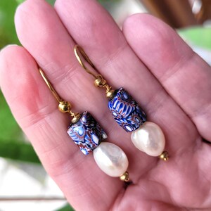 May include: A pair of drop earrings featuring gold-tone hooks, small gold beads, cylindrical millefiori glass beads with blue, white, and reddish-brown floral patterns, and dangling white baroque-style pearls at the bottom of each earring.