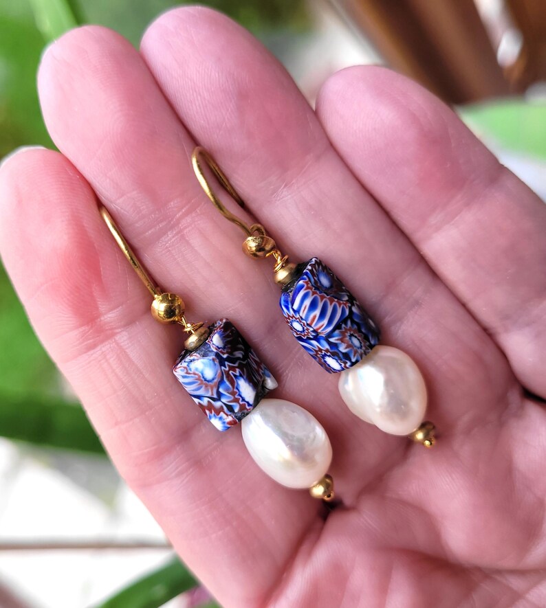 May include: A pair of drop earrings featuring gold-tone hooks, small gold beads, cylindrical millefiori glass beads with blue, white, and reddish-brown floral patterns, and dangling white baroque-style pearls at the bottom of each earring.
