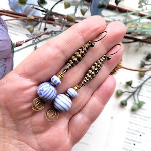 May include: A pair of long dangle earrings featuring antique brass-toned metal components with a textured, beaded design. Each earring includes two round, white ceramic beads with blue horizontal stripes and a flat, coiled brass wire accent at the base.