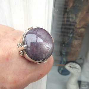 Puede incluir: Un anillo grande, de forma ovalada, con una piedra preciosa pulida de color gris violáceo. El engaste del anillo es plateado, con detalles decorativos en los laterales. La piedra preciosa refleja la luz, creando un efecto brillante. Una joya llamativa.
