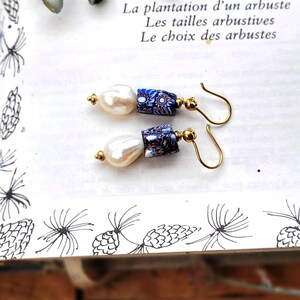 May include: A pair of drop earrings featuring white baroque pearls paired with cylindrical beads in a blue, white, and red floral millefiori pattern. The components are connected by gold-toned metal findings and attached to gold-toned curved ear wires.