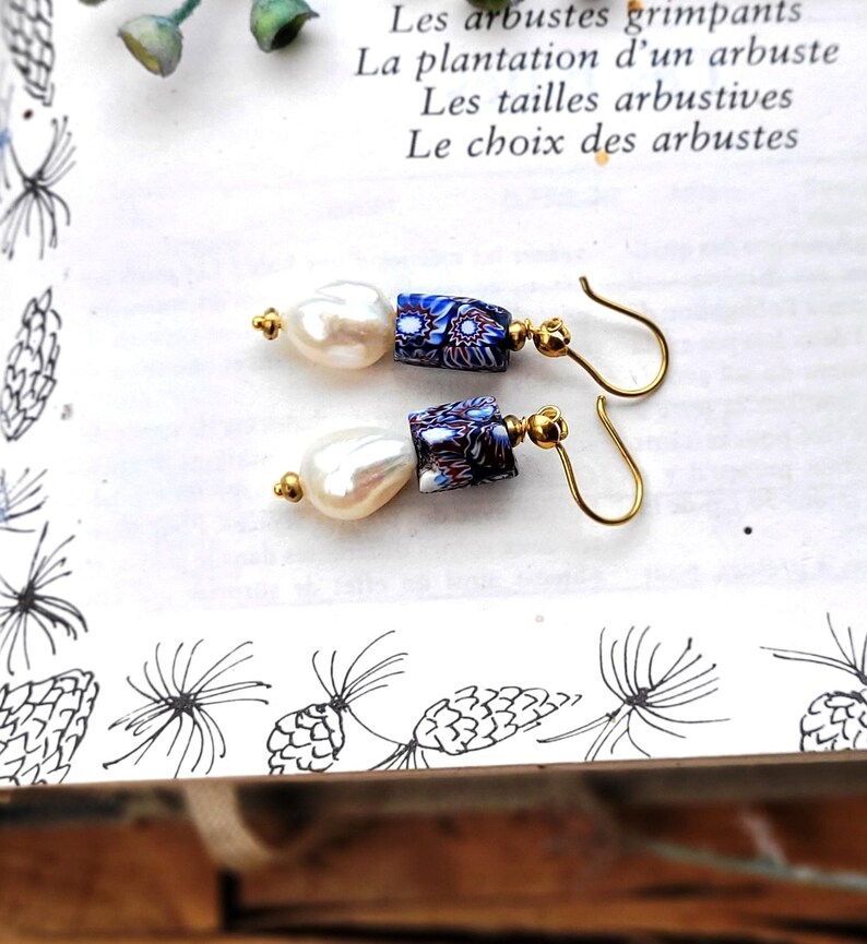May include: A pair of drop earrings featuring white baroque pearls paired with cylindrical beads in a blue, white, and red floral millefiori pattern. The components are connected by gold-toned metal findings and attached to gold-toned curved ear wires.